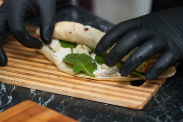 Man's Hands Rolling Russian Thinny Pancakes with Cheese Cream and Spinach on Cutting board over Kitchen Table.