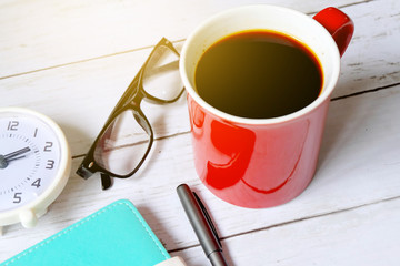 Selective focus and top view of black coffee in red mug with notebook,pen ,clock and sunglasses on white wooden background.