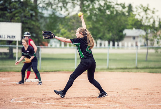 A teenaged girl in baseball pitching motion on a pitchers mound with long hair in a ponytail and black and green ball uniform - Powered by Adobe