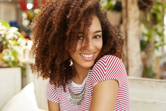 Half length portrait of beautiful attractive African American woman sits against cafe interior, has shining smile on face, being in high spirit after walk with boyfriend outdoor. Positive emotions