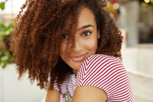 Sideways portrait of stylish female with curly Afro hairstyle and dark skin, being satisfied to achieve great success in life, poses indoor. Close up shot of beautiful African woman has free time