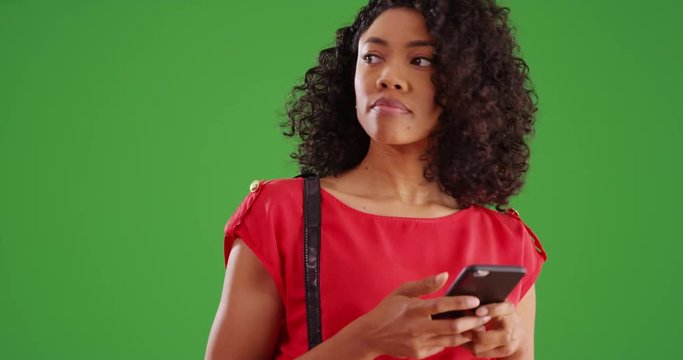 Pretty Black woman texting with cell phone and smiling on greenscreen background. African American Millennial using messaging app on smartphone and looking around her on green screen. 4k