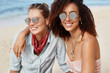 Lesbian couple spend summer vacations at tropical beach, embrace each other and enjoy togetherness. African American female in shades has homosexual relationships with Caucasian blonde woman