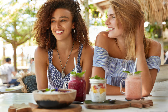 Pleased female lesbians meet together at cafe, enjoy tasty desserts, have recreation at tropical country, discuss something positive. Mixed race women sit against outdoor terrace cafeteria interior