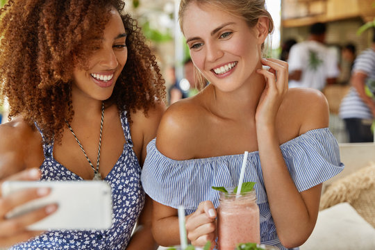 Cropped shot of cheerful young feminists pose against cafe interior with smoothie, make selfie on modern mobile phone or talk online on video call, share their impressions about resort abroad