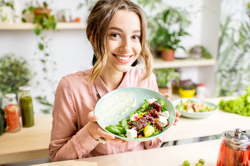 Portrait of a young woman holding a plate of salad sitting indoors surrounded with green flowers...