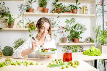 Young woman eating healthy food sitting in the beautiful interior with green flowers on the background