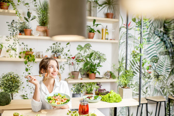 Young woman eating healthy food sitting in the beautiful interior with green flowers on the...