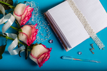 on a blue background lie three fragile pink roses with a white ribbon, scattered beads, lies an open blank notebook, a colored pen