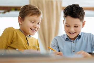 Having fun. Cheerful pleasant pre-teen boys sitting at the table and drawing together while smiling and discussing the picture