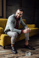 fashionable smiling man with cigar and ashtray sitting on couch