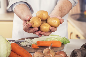 A man holds potatoes in his hands over a kitchen table with vegetables for cooking a vegetarian dish.