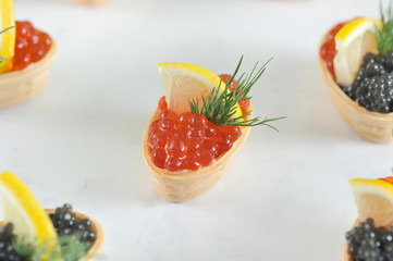Basket with red caviar in the center of the frame. The appetizer is decorated with dill and a slice of lemon. The edges of the basket with red and black caviar. Light background. View from above.