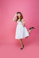 Adorable caucasian woman in white dress taking off sunglasses and looking aside on copyspace with surprise, isolated over pink background