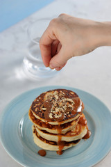 Pancakes are laid one on another, are poured with caramel sauce and sprinkled with grated almonds. Over the pancakes is brought a hand, sprinkled with pancakes nuts. Vertical shot.
