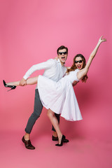 Full-length amazing photo of beautiful dancing couple in sunglasses enjoying party together, isolated over pink background