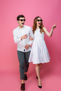 Full-length image of luxury lifestyle man and woman in sunglasses walking together on camera, isolated over pink background