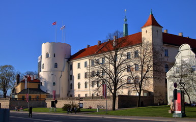 Zamek w Rydze, stolicy Łotwy, mały park przed budowlą, ludzie spacerują, siedzą na ławkach i na trawie, słoneczny dzień, błękitne bezchmurne niebo - obrazy, fototapety, plakaty