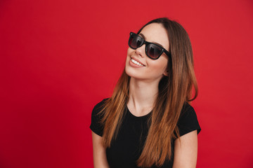 Closeup photo of fashion woman 20s wearing black t-shirt and sunglasses looking on camera with perfect smile isolated over red background