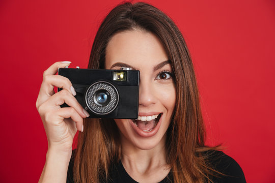Image of happy professional photographer wearing black t-shirt holding retro camera at face and making photos, isolated over red background