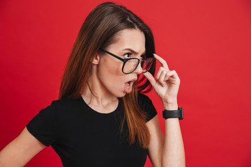 Photo of young outrageous woman wearing black t-shirt taking off glasses and looking aside with indignation or shock isolated over red background