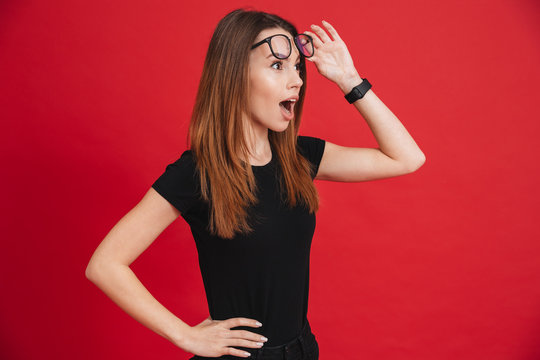 Closeup image of young woman 20s wearing black t-shirt taking off glasses and looking aside in surprise isolated over red background