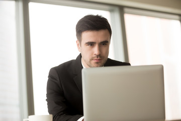 Portrait of handsome millennial businessman working on laptop, successful CEO reading business news online, using computer in office. Trader or broker monitoring exchange rate fluctuations in Internet
