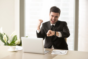 Excited businessman yelling with joy when sitting at desk in front of laptop and looking at wrist...