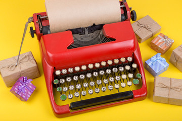 Holidays concept - red typewriter with blank craft paper, gift boxes on yellow background
