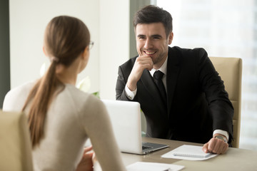 Handsome smiling male office worker in business suit sitting at desk with laptop in front of female...