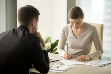 Satisfied with loan terms woman singing contract when sitting at desk in front of male bank employee at office. Millennial female applicant accepting employers offer and putting signature in agreement