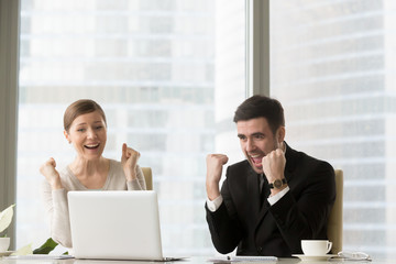 Happy female and male managers or business partners screaming with joy when sitting at desk and looking on laptop screen. Young businessman and businesswoman enjoying work results, celebrating victory