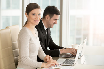 Portrait of beautiful young woman sitting at desk in front of laptop together with male colleague and looking at camera with smile. Female and male company managers working on computers at workplace
