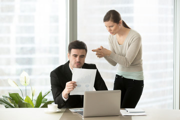Angry woman aiming with finer gun at head of her hateful boss who attentively reading financial report at desk. Intrigues of office colleagues, bad coworkers relations, hidden negative among employees