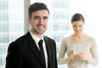 Portrait of handsome smiling millennial businessman in black suit standing in office, female secretary using digital tablet on background. Successful and confident company leader looking at camera