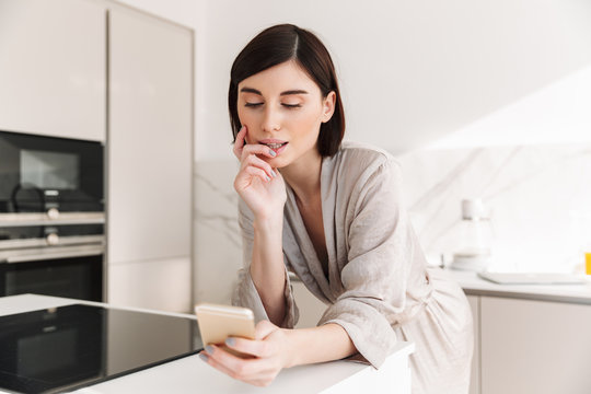 Photo of attractive woman with short dark hair wearing beautiful robe posing in kitchen, and chatting or surching internet on smartphone