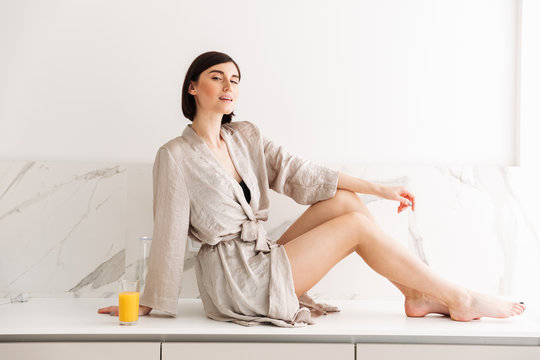 Portrait of sexual slim woman with short dark hair wearing robe posing in kitchen, sitting on table in sunny morning and drinking orange juice