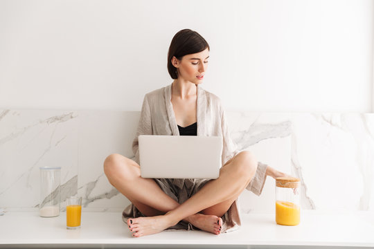 Image of smart brunette woman wearing bathrobe sitting on table in kitchen with legs crossed, and using laptop