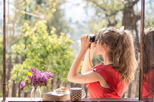 Young girl with vintage red dress watching birds in the morning sun by the window, while tea , cookies , book and lilac flowers are waiting next to her