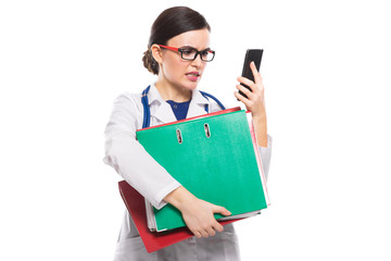 Angry young woman doctor with stethoscope holding binders and phone in her hands in white uniform on white background