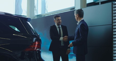 Side view of manager in suit shake hand to adult businessman in showroom near black car.