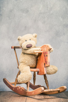 Teddy Bear toy sitting on old retro wooden rocking horse front concrete wall background. Vintage style filtered photo