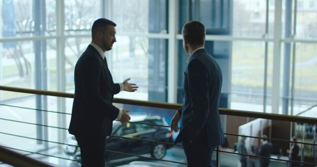 Two elegant men having conversation on balcony in car showroom.