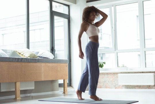 Full length cheerful woman with curly hair standing on mat while taking exercise in apartment
