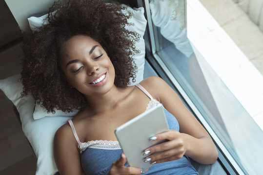 Portrait of outgoing african woman looking at sreen of electronic tablet while lying on comfortable bed in room. Top view