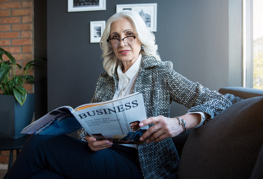 Reading news. Portrait of stylish attractive optimistic mature businesswoman in glasses is holding journal and looking at camera with slight smile while sitting on couch in office