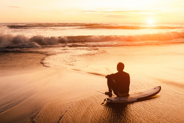 surfer sits on his board near the sea at sunset - obrazy, fototapety, plakaty