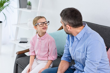 Obraz premium father and daughter in eyeglasses looking at each other while sitting on sofa at home