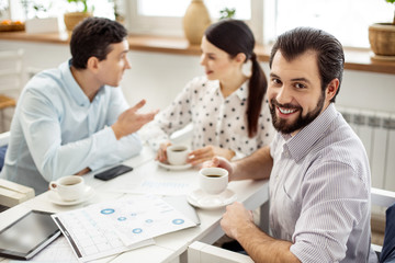 Working day. Good-looking cheerful bearded man drinking coffee and smiling and his colleagues talking in the background