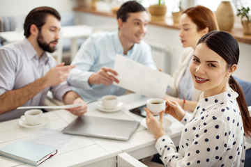 Fototapeta premium Working atmosphere. Attractive alert dark-haired young woman smiling and drinking coffee and her colleagues talking in the background
